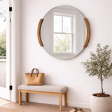 Round mirror with wooden frame on a white wall above a bench with a bag, next to a plant and shoes on a beige rug.