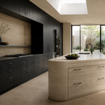 A modern kitchen interior with a curved light beige stone island featuring a white countertop and dark wood cabinetry along one wall. Large windows and a skylight provide natural light, and an outdoor garden is visible in the background.