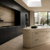 A modern kitchen interior with a curved light beige stone island featuring a white countertop and dark wood cabinetry along one wall. Large windows and a skylight provide natural light, and an outdoor garden is visible in the background.